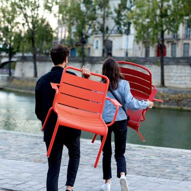 Bright coloured aluminium chairs being carried by a woman and a man by a Paris river