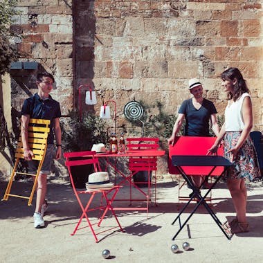 Three people set out bright Fermob Bistro folding square tables and chairs in the sun for a game of boules