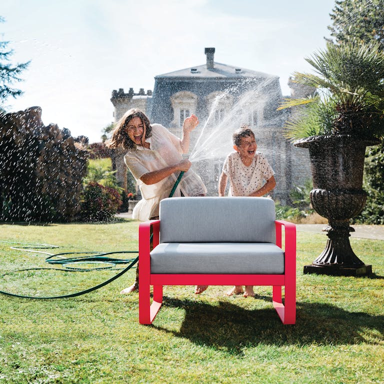 Woman sprays hose over boy in garden and gets Pink Praline Fermob Bellevie sofa chair wet
