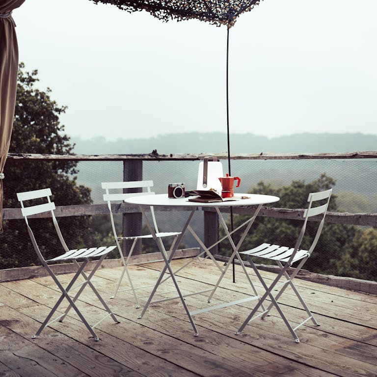 A Fermob Bistro 96cm metal folding round table and chairs in Cotton White sit on a rustic deck