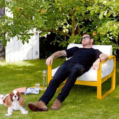 Man relaxes on Honey coloured Fermob Bellevie sofa armchair in a garden with dog