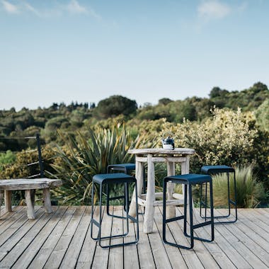 Four Fermob Bellevie contemporary bar stools sit around a rustic table on a deck overlooking bush